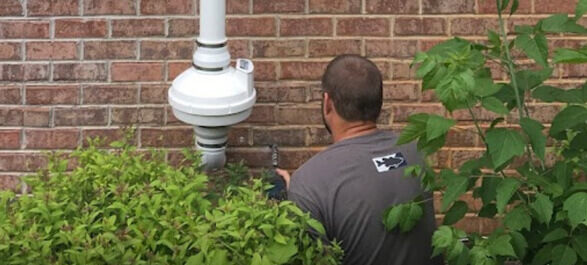 The image shows an expert performing radon mitigation system installation on a home’s exterior wall. The technician is installing the radon vent pipe connected to the radon mitigation system designed to reduce harmful radon gas levels within the home. Radon gas, a naturally occurring radioactive substance, is a potential health risk when it accumulates indoors, and this system is vital for improving indoor air quality and reducing radon exposure. The expert is ensuring the system is properly designed, installed, and functioning to meet safety and health standards.